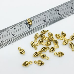 以下が含まれることがあります： 小さな金色の松ぼっくりチャーム。ジュエリー作りのためのループが付いています。背景にはセンチメートル単位の金属製の定規があります。チャームは白い表面に散らばっています。