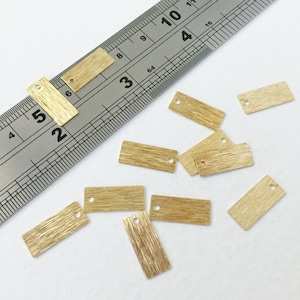 May include: Several small, rectangular brass tags with a wood grain effect and a single hole. A metal ruler with centimetre measurements is in the background. The tags are scattered on a white surface.