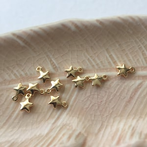 Könnte beinhalten: Eine Gruppe goldener sternförmiger Charms mit Sprungringen. Die Charms sind auf einer hellbraunen Oberfläche angeordnet.
