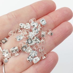 May include: A pile of silver earring backs, also known as earring clutches, in a hand. The earring backs are made of metal and have a small loop on one side and a flat back on the other side.