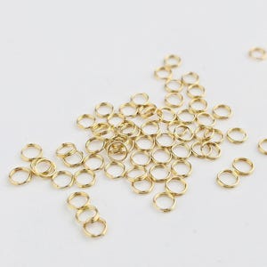 May include: A collection of small, gold-colored split rings scattered on a white surface. These metal rings are circular with a split, allowing them to be opened and attached to other objects. The rings are likely used for jewelry making or crafts.