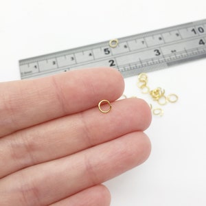 May include: A close-up of a gold jump ring, approximately 5 mm in diameter, held in a hand. A ruler is visible in the background, with a measurement of 5 cm.