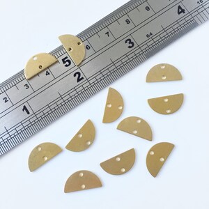 May include: Ten brass half-circle shapes with two holes each. The shapes are scattered on a white surface. A ruler is in the background.