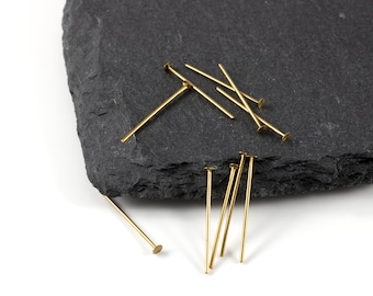 May include: A group of gold-colored head pins with a flat head on a black slate surface. The pins are arranged in a random pattern.