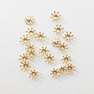 May include: A collection of small, gold-colored metal bead caps, each with a starburst design. The caps have a central hole and multiple rounded points extending outwards, scattered on a white surface. These are likely used for jewelry making or crafting.