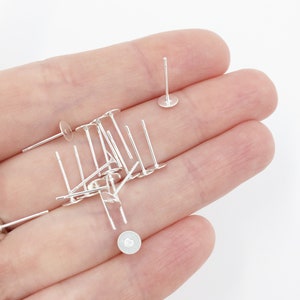 May include: A close-up of a hand holding a pile of silver earring posts. The posts are approximately 2.5 cm long and have a small, round pad at the bottom.