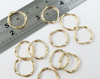 May include: Gold-coloured, twisted metal rings scattered on a white surface. A metal ruler with black markings is in the background. The rings have a wavy, circular design, and are likely jewellery components or craft supplies.
