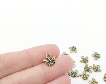 May include: A close-up of a gold-toned metal bee charm with a loop for attaching to jewelry or crafts. The charm is about 1 cm in size and has a detailed design.