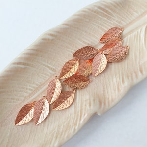 May include: A collection of rose gold leaf charms with a hole at the top for stringing or attaching to jewellery. The charms are arranged in a cascading pattern on a light beige surface.