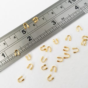 May include: A close-up of a ruler measuring 28 cm with a row of 18 gold-coloured metal crimp beads scattered on a white surface.