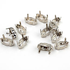May include: A collection of silver-colored, oval-shaped jewelry settings. Each setting has four prongs and a base with small posts. The settings are scattered on a white surface, showcasing their design and potential use in jewelry making.