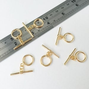 以下が含まれることがあります： 丸い輪とストレートバーが付いた、金色の金属製のトグル留め具が5つ。留め具は白い表面に配置され、背景には定規があります。