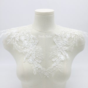 May include: White lace applique with floral designs and beaded accents. The applique is displayed on a white mannequin bust. "Beads Factor" is written in the center of the applique.