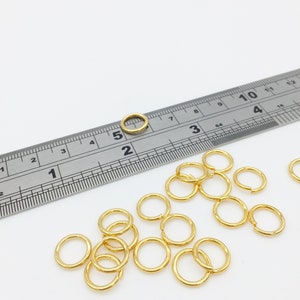 以下が含まれることがあります： 定規で18個の金色のジャンプリングの山を測定している様子のクローズアップ。ジャンプリングの直径は約5mmです。