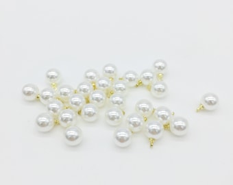 May include: A group of white pearl beads with gold accents. The beads are scattered on a white surface.