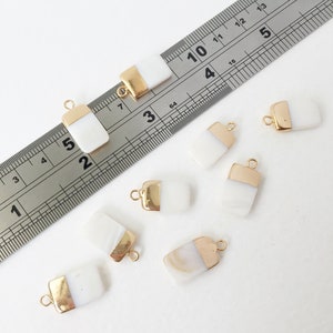May include: Seven white and gold rectangular pendant charms with a gold loop at the top. The charms are approximately 2.5 cm long and 1.25 cm wide. The charms are arranged on a white surface with a ruler in the background.