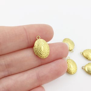 以下が含まれることがあります： 金色の楕円形ロケットペンダント。手のひらに乗せられており、背景にもいくつか見えます。チェーンやブレスレットに取り付けるための小さなループが付いています。ロケットペンダントの長さは約2.5cmです。