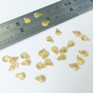 以下が含まれることがあります： 小さな金色の貝殻チャームのコレクション。各チャームには、ジュエリーやその他の工芸品に取り付けるためのループが付いています。背景には、センチメートル単位の目盛りが付いた金属製の定規があります。