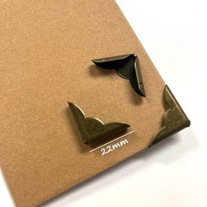 May include: Three antique bronze-coloured metal corner protectors attached to a tan rectangular card. The corner protectors feature a decorative scalloped edge design. A white line and the text 22mm indicate the size of the metal components.