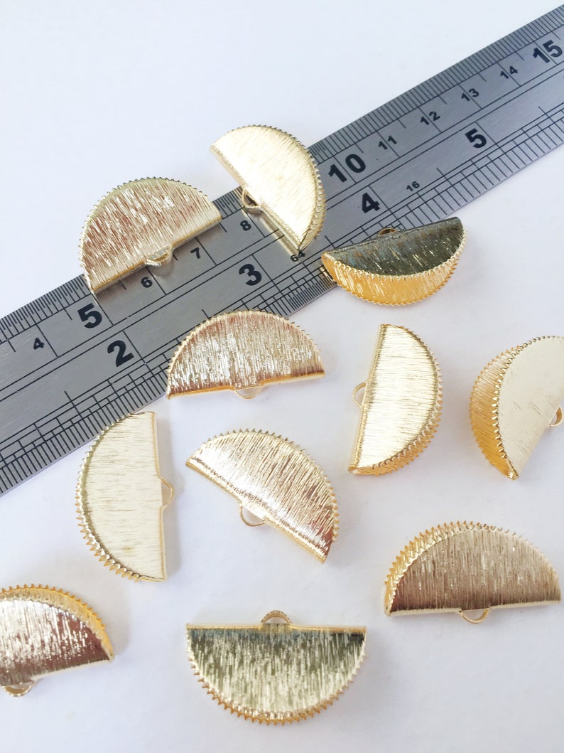 May include: Ten gold-coloured metal half-circle embellishments with a textured surface and a small loop on the top. The embellishments are scattered on a white surface.