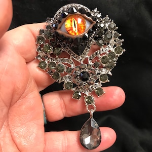 May include: A silver-toned brooch featuring a central eye with orange and yellow iris, surrounded by dark gray and clear crystals. A teardrop-shaped crystal hangs from the bottom. The brooch has an ornate, floral design.
