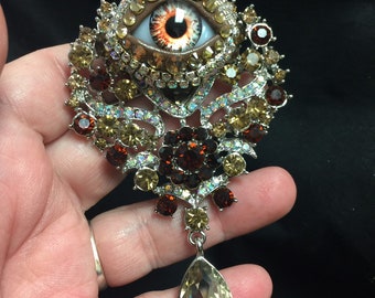 May include: A silver brooch with a large, brown eye surrounded by rhinestones. The brooch has a teardrop-shaped pendant with a clear rhinestone.