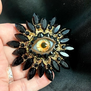 Könnte beinhalten: Eine große, schwarze und goldene Augenbrosche mit brauner Iris und schwarzer Pupille. Das Auge ist von einem Ring aus schwarzen Strasssteinen und einem goldenen Filigranmuster umgeben. Die Brosche ist auf einem schwarzen Stoffhintergrund befestigt.