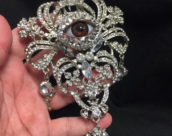 Peut inclure: Une broche en argent et en cristal avec un grand œil réaliste au centre. L'œil est brun avec un iris blanc et une pupille noire. La broche est décorée de nombreux petits cristaux.
