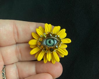 Peut inclure: Une broche en forme de tournesol jaune avec un motif d'œil central. L'œil est bleu clair avec une pupille foncée, entouré de pierres précieuses marron et dorées. La broche est tenue dans une main sur fond noir.