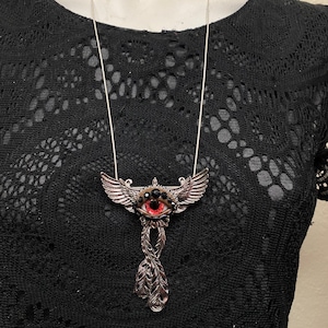 May include: A silver-toned pendant necklace featuring a central red eye with black accents, flanked by stylized wings. The pendant has a feather-like design below the eye. The necklace is displayed against a black lace top.