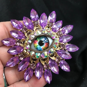 Può includere: Un grande occhio colorato con un'iride arcobaleno è circondato da un cerchio di strass trasparenti e un anello di cristalli marroni a forma di marquise.