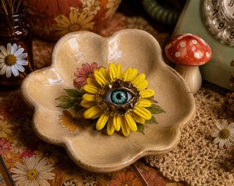 Könnte beinhalten: Eine gelbe, sonnenblumenförmige Brosche mit einem realistischen blauen Auge in der Mitte, das von kleinen bernsteinfarbenen Kristallen umgeben ist. Die Brosche liegt in einer beigen, blumenförmigen Keramikschale mit einem floralen Muster.