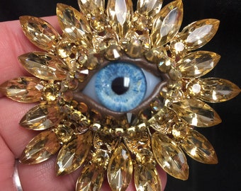 Peut inclure: Un grand œil bleu avec un iris brun, entouré d'un motif de fleur dorée fait de strass.