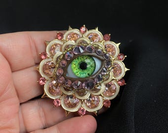 Peut inclure: Broche détaillée avec un œil central à l'iris vert vif, entouré d'un anneau de cristaux violets et roses. L'œil est serti dans un cadre doré en forme de fleur, orné de cristaux roses supplémentaires.