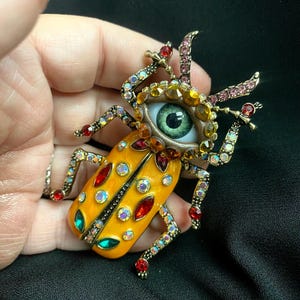 Könnte beinhalten: Ein dekorativer Käfer-Broche mit einem gelben Körper, roten und grünen Akzenten und schillernden Kristallen. Die Brosche hat ein zentrales Auge mit grüner Iris und kristallbesetzte Beine und Fühler. Ein einzigartiges Schmuckstück.