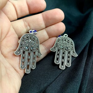 Może przedstawiać: Dwa srebrne metalowe wisiorki w kształcie ręki Hamsa ze skomplikowanymi wzorami filigranowymi i małymi okrągłymi wycięciami. Każdy wisiorek ma mały niebiesko-biały koralik w kształcie oka przymocowany do g&oacute;rnego k&oacute;łeczka z ażurowym wzorem.