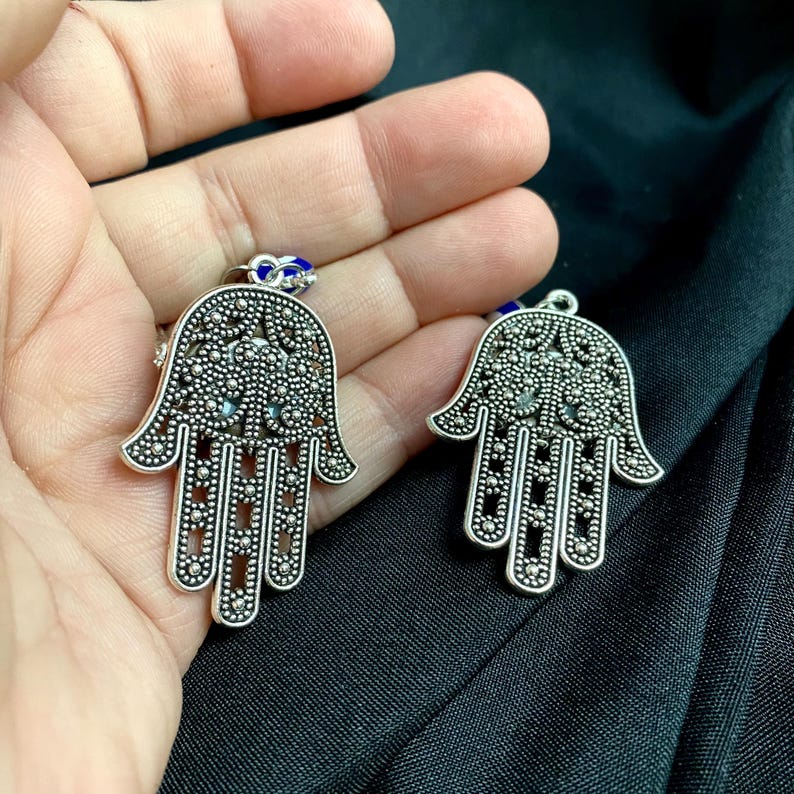 Może przedstawiać: Dwa srebrne metalowe wisiorki w kształcie ręki Hamsa ze skomplikowanymi wzorami filigranowymi i małymi okrągłymi wycięciami. Każdy wisiorek ma mały niebiesko-biały koralik w kształcie oka przymocowany do g&oacute;rnego k&oacute;łeczka z ażurowym wzorem.