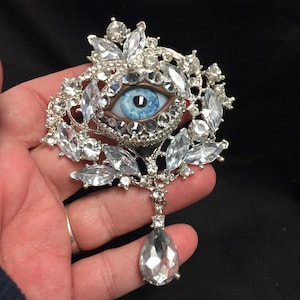 Könnte beinhalten: Silberfarbene Brosche mit einem zentralen Augendesign, das eine blaue Iris und einen braunen Rand aufweist. Umgeben von klaren, funkelnden Kristallen, mit einem tropfenförmigen Kristallanhänger.