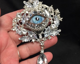 Peut inclure: Broche argentée ornée d'un œil central avec un iris bleu et un contour marron. Entouré de cristaux transparents étincelants, le bijou comporte un pendentif en cristal en forme de goutte.