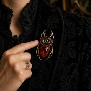May include: A beetle-shaped brooch featuring a central green eye surrounded by deep red faceted stones and clear crystals on the pincers. The insect body is crafted with gold-toned metal legs and a large, teardrop-shaped red gemstone at the base.