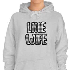 May include: A light gray hoodie with the words "LINE WIFE" in black and white buffalo plaid lettering. The hoodie has a drawstring hood and a front pocket. The model is wearing dark blue jeans.
