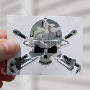 May include: A white sticker with a skull wearing a camouflage helmet and crossed wrenches. The skull is in a camouflage pattern.