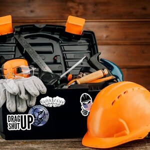 May include: A black toolbox filled with tools, including a level, a hammer, safety glasses, ear protection, and work gloves. The toolbox has stickers on it, including one that says "Drag Up Shut Up". An orange hard hat sits next to the toolbox.