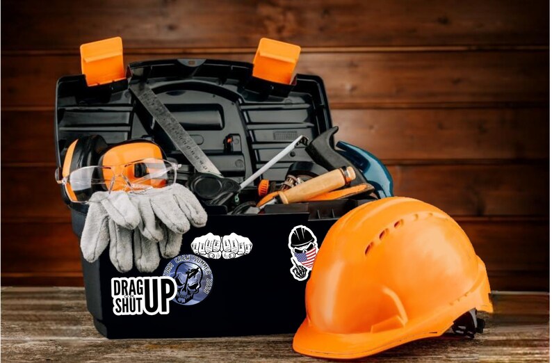 May include: A black toolbox filled with tools, including a level, a hammer, safety glasses, ear protection, and work gloves. The toolbox has stickers on it, including one that says "Drag Up Shut Up". An orange hard hat sits next to the toolbox.