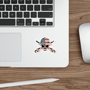 May include: A sticker with a skull wearing a hard hat with the American flag on it. The skull is holding two crossed wrenches.