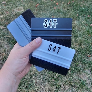 Puede incluir: Dos rasquetas negras y blancas con un borde de fieltro negro y un mango de plástico gris. Las rasquetas tienen una pegatina blanca con el texto "S4T" en ellas.