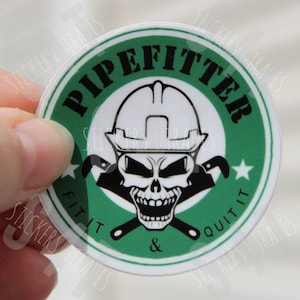 May include: A round sticker with a green background and white border. The sticker features a skull wearing a hard hat and holding two wrenches. The text "PIPEFITTER" is written in a black circle around the skull. The text "FIT IT & QUIT IT" is written in a black circle around the skull.