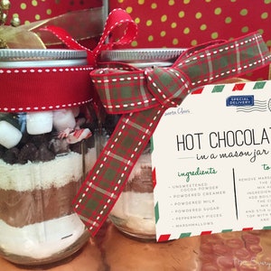 May include: Two mason jars filled with hot chocolate mix ingredients, including cocoa powder, powdered creamer, milk, sugar, peppermint pieces, and marshmallows. A red and green plaid ribbon is tied around each jar. A white card with the text "HOT CHOCOLATE in a mason jar" and a list of ingredients and instructions is attached to the jars.