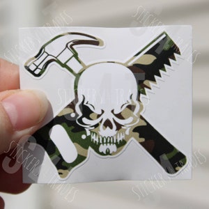 May include: White skull with a hammer and saw crossed behind it. The tools are in a camouflage pattern.
