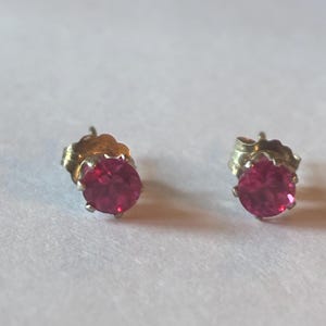 May include: A pair of stud earrings featuring round, bright red gemstones set in gold-colored metal. The earrings are displayed on a white surface, showcasing their simple yet elegant design. The gemstones have a vibrant, eye-catching color.